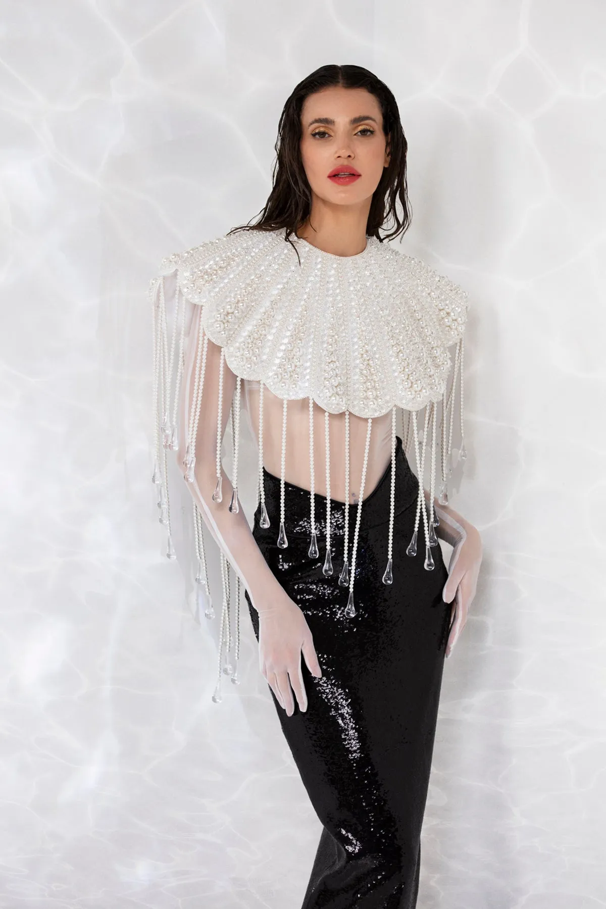 Scallop shell inspired ensemble consisting of a cropped top embroidered with pearls, crystals, and hand blown glass drops. Worn with a white sheer bodysuit and a pencil skirt in high gloss sequins